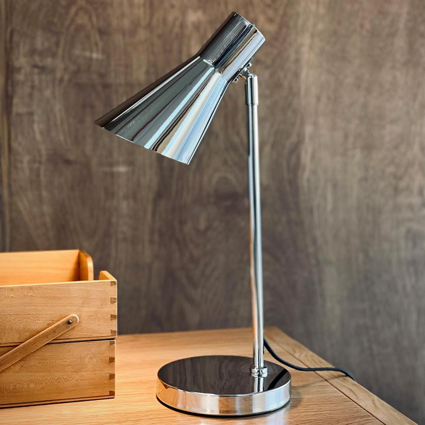 A chrome desk lamp points down towards the tabletop of a light wooden table; a wooden box stands on the left in front of a dark wood-clad wall.