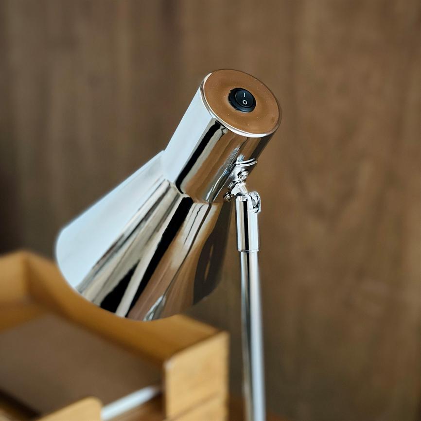 Chrome desk lamp stands at an angle with visible on/off button; its glossy screen reflects the surroundings in front of a blurred wooden background and a small wooden box.