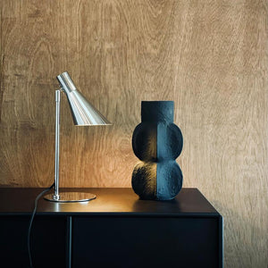 Desk lamp shines dimly against a sculptural, black vase; both stand on a dark cabinet in front of a warm, wood-clad wall.