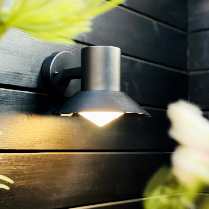Outdoor wall lamp shines downwards against black wooden panels and casts a warm, low glow; blurred leaves in the foreground suggest a nearby garden or terrace.