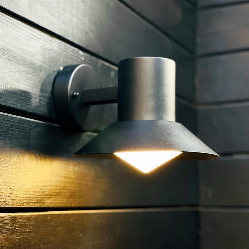 A matte black wall lamp casts a warm downward light, casts a triangular cone of light and is mounted on horizontal dark wood panels on an outdoor or covered wall.