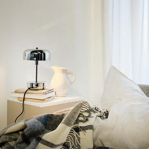 Chrome table lamp stands on a bedside table next to a white jug; a stack of books lies under the lamp; striped pillow and checkered plaid form a cosy, bright bedroom nook.
