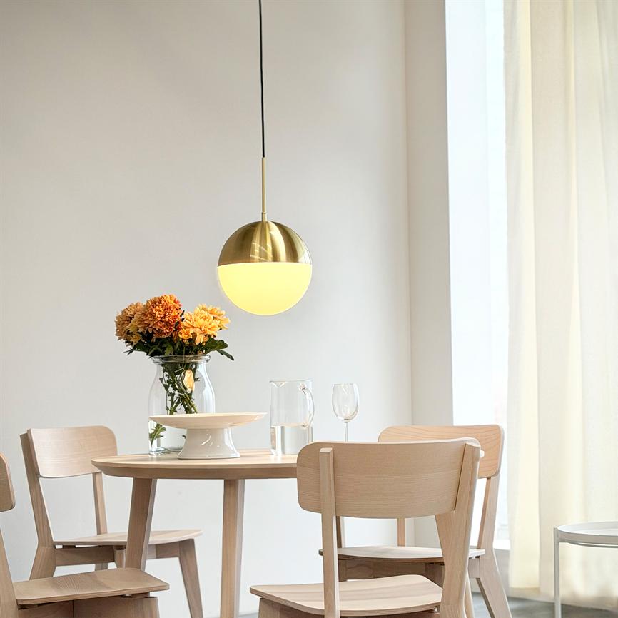 A round golden pendant light shines above a round wooden table with four light wooden chairs; the table has a glass vase with orange flowers, a decanter, a wine glass and light curtains in the background.
