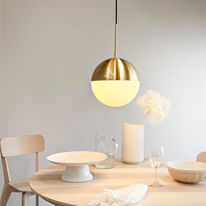 A gold-plated pendant lamp hangs and casts warm light over a bright wooden table with a porcelain dish, glass jug, wine glass, bowl, vase with dried flowers and two wooden chairs.