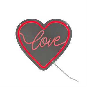 Heart-shaped neon sign (text: "love") shows red neon light along the edge and handwritten "love" in the middle, mounted on gray plate with power cord and two hanging holes.
