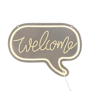 The neon sign "welcome" glows warm yellow; it is shaped like a speech bubble in a grey-brown plate with a neon edge, hanging holes and white wire on a white background.