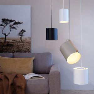 Pendant lamps hang at heights; a beige lamp shines downwards. Next to it is a gray sofa with an open book, a brown plaid and a yellow ribbed pillow in front of a large sun patch of trees.