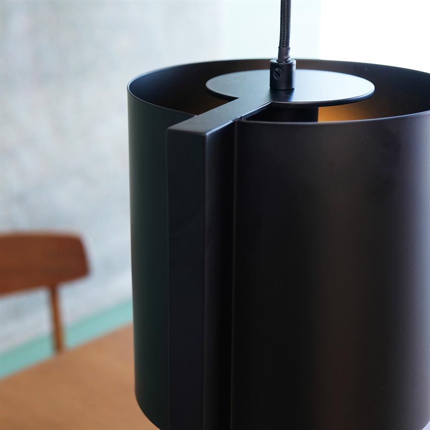 A black cylindrical pendant lamp hangs above a wooden table; the lamp's top metal rod and cord are visible, with a blurred wooden chair and a light, textured wall in the background.