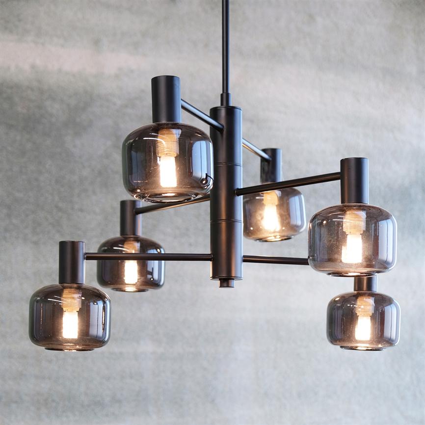 A modern ceiling lamp with six smoky glass domes, where the bulbs glow weakly, suspended in black metal arms in front of a textured light gray wall.