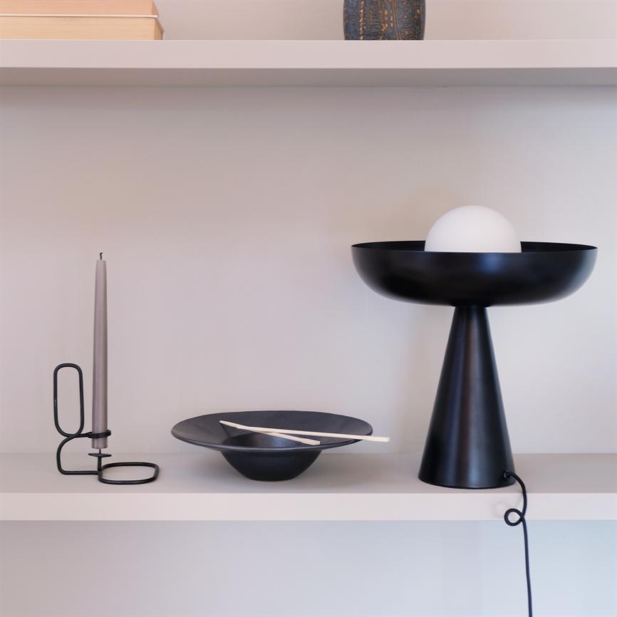 Black conical table lamp with white glass ball is on the right; in the middle of the shelf rests a black bowl with two bright chopsticks; on the left a gray candle in a black metal holder. White wall.