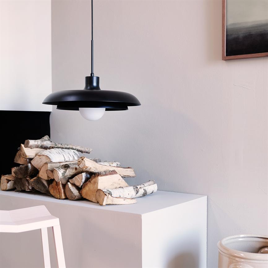 Black hanging pendant lamp shines above a stack of birch logs on a white shelf in a bright, minimalist room with a bar stool and a ceramic jar.