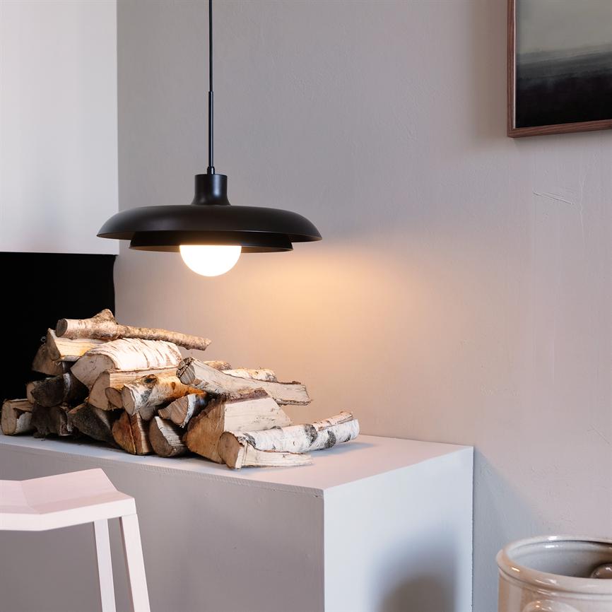 Black pendant lamp hangs and shines on a stack of birch wood pieces placed on a white shelf, in a quiet, minimalist room with light walls and a dark art frame in the corner.