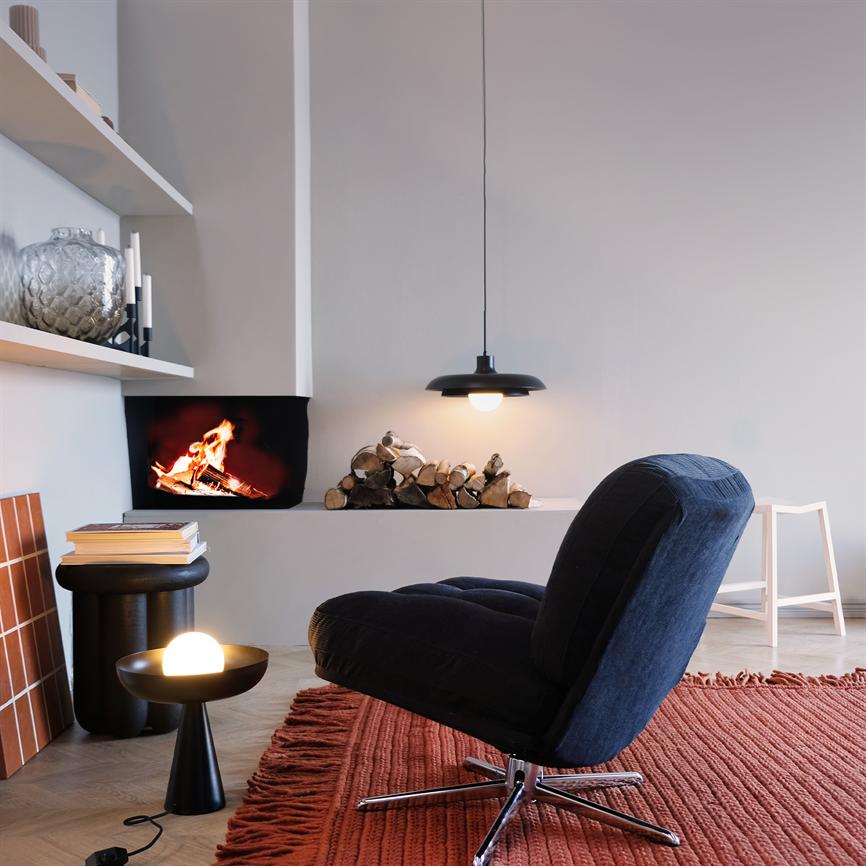 A black lounge chair faces a fire in the fireplace in a modern, minimalist living room with a stack of firewood, a hanging lamp, a small table with a round lamp and a red carpet with fringes.