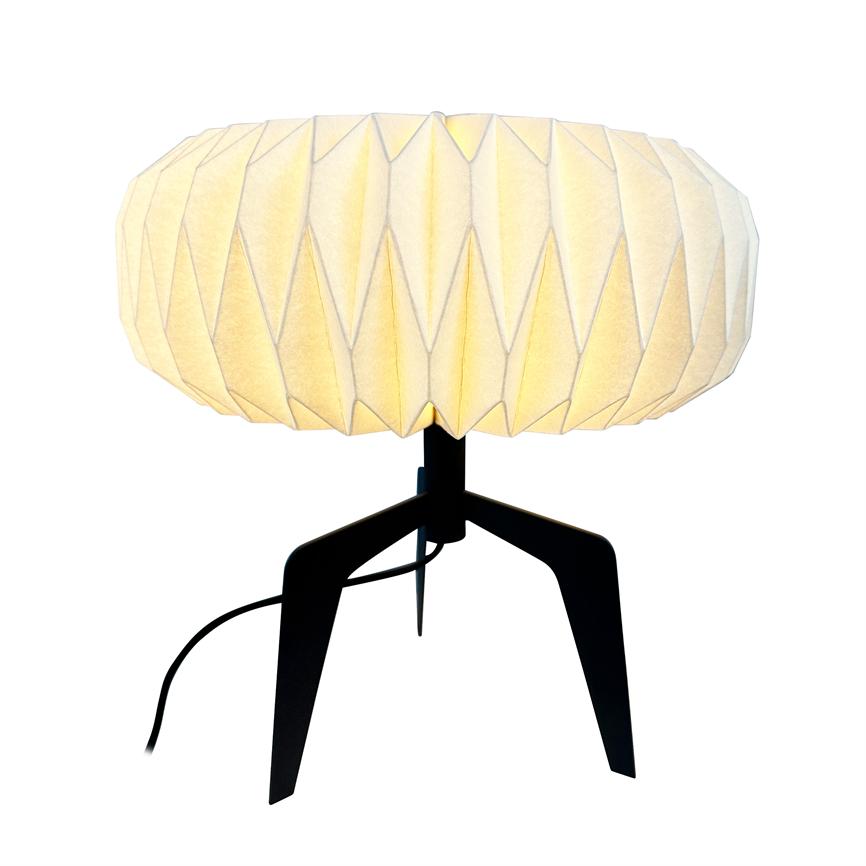 Table lamp with a round, folded paper shade that emits a warm, diffused light; stands on black tripod with visible wire, isolated against a white background.