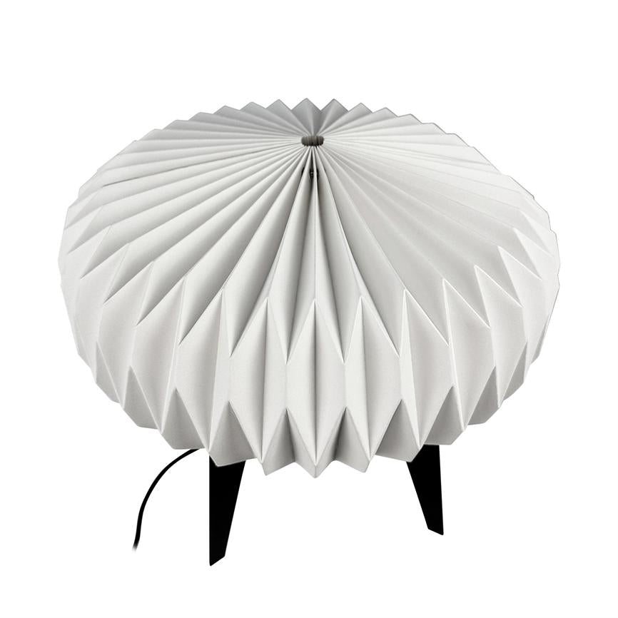 Round lamp with pleated white shade rests on three black legs; on a pure white background, a black power cable can be seen running from the lower edge of the screen.
