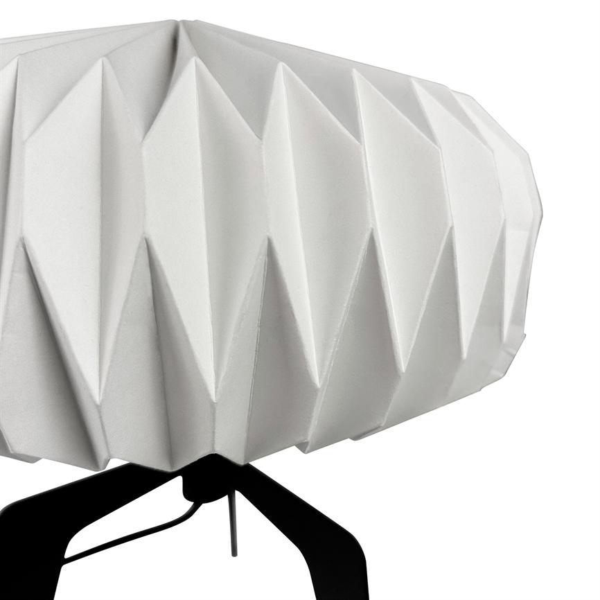 A white, folded lampshade shows sharp, triangular folds; it sits on a black three-legged stand against a neutral white background.