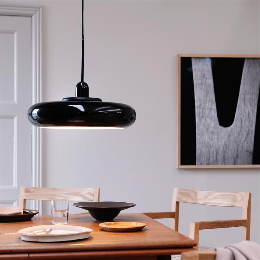 Black pendant lamp hangs above a wooden dining table; ceramic dishes, a water glass and light wooden chairs stand ready in a bright, minimalist dining room with an abstract black-and-white picture on the wall.