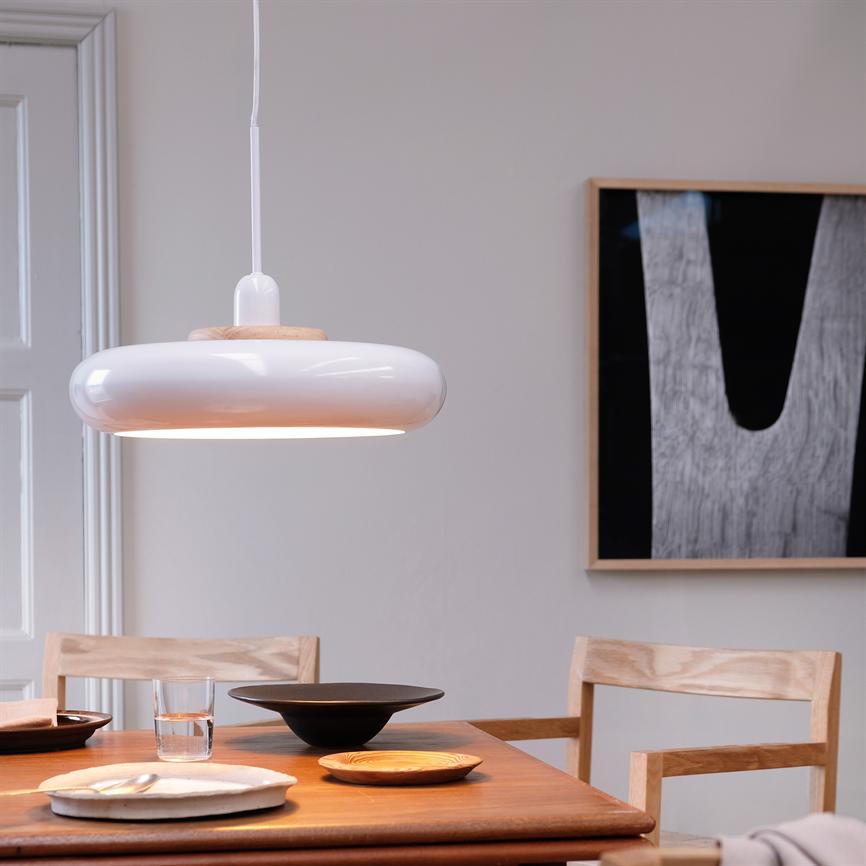 A white pendant lamp hangs and casts warm light over a wooden dining table with plates, glasses and wooden chairs in a bright room with a black and white framed painting on the wall.