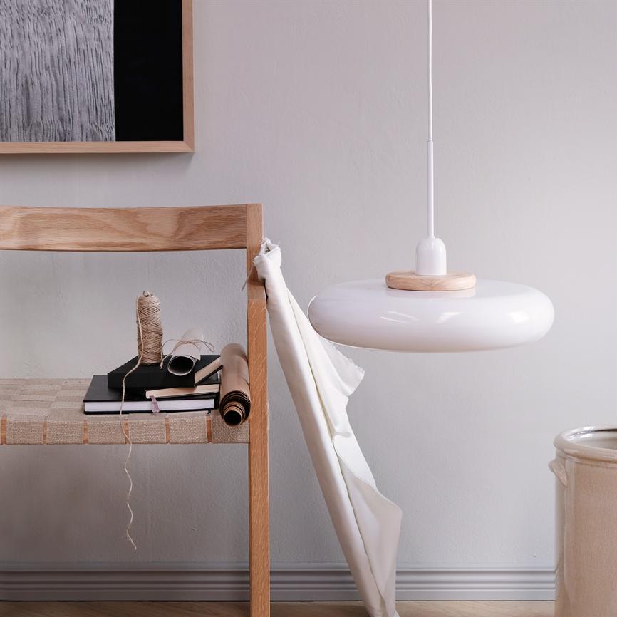 White round pendant lamp hangs above a light wooden chair; the chair carries rolled up papers, books and a spool of string, while a piece of white fabric hangs down against a light gray wall.