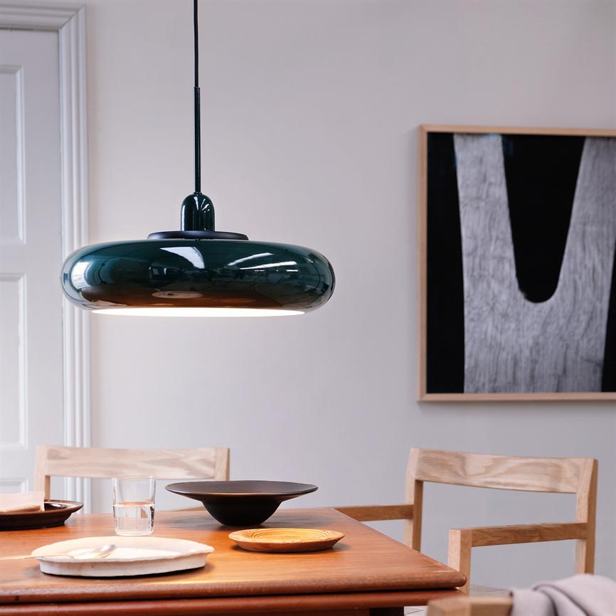 A dark green glazed pendant light down over a wooden table with plates, bowls and a glass; surrounded by light wooden chairs and abstract black and white wall art in a bright, minimalist space.