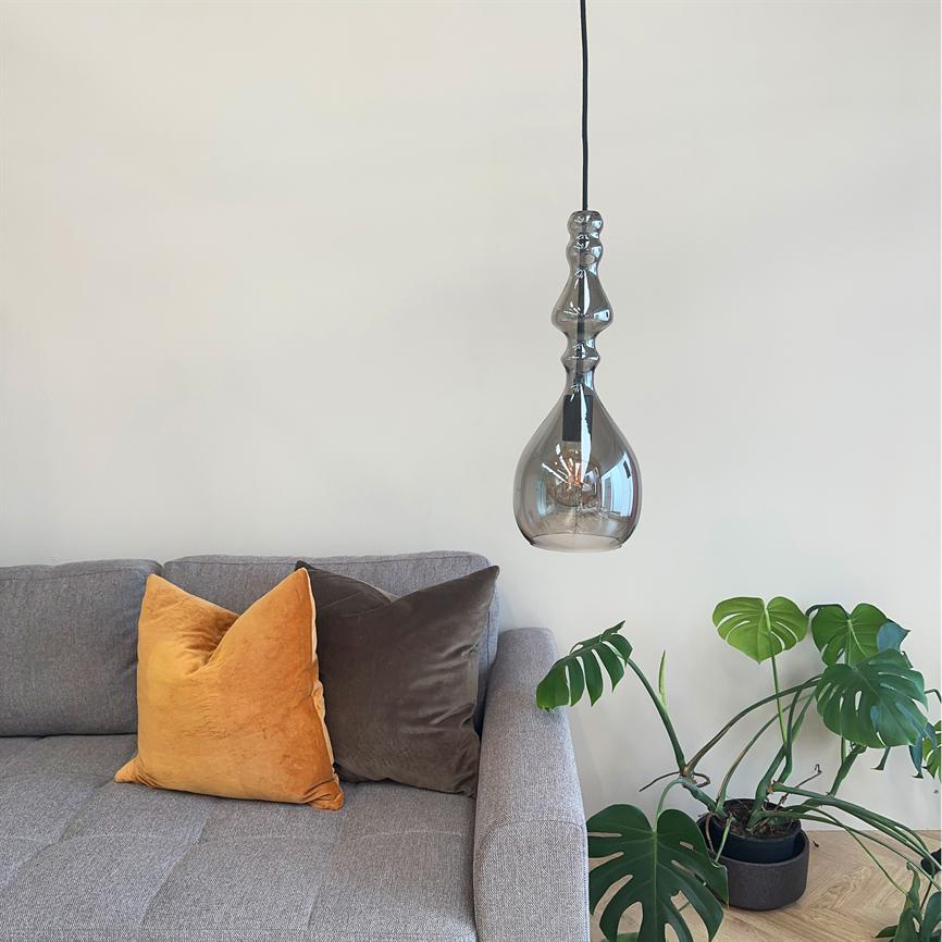 A decorative hanging glass bulb floats above a gray sofa; two velor cushions (yellow and brown) lie on the sofa, and a large potted monstera plant stands on the wooden floor.