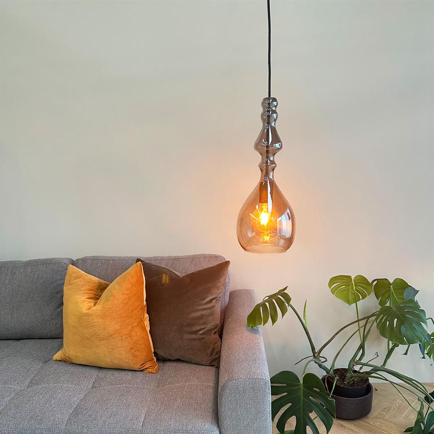 Hanging glass bulb glows warmly above a gray sofa; two decorative pillows (orange and brown) lie on the sofa, and a green monstera plant stands in a pot on the wooden floor.