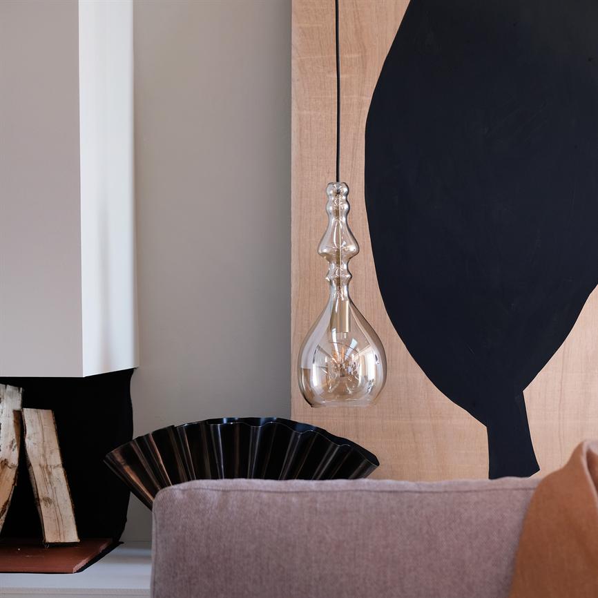 Glass pendant hangs in front of a light wood panel with black abstract form; an upholstered sofa in the foreground, a black decorative bowl and stacks of firewood by a modern fireplace in the background.