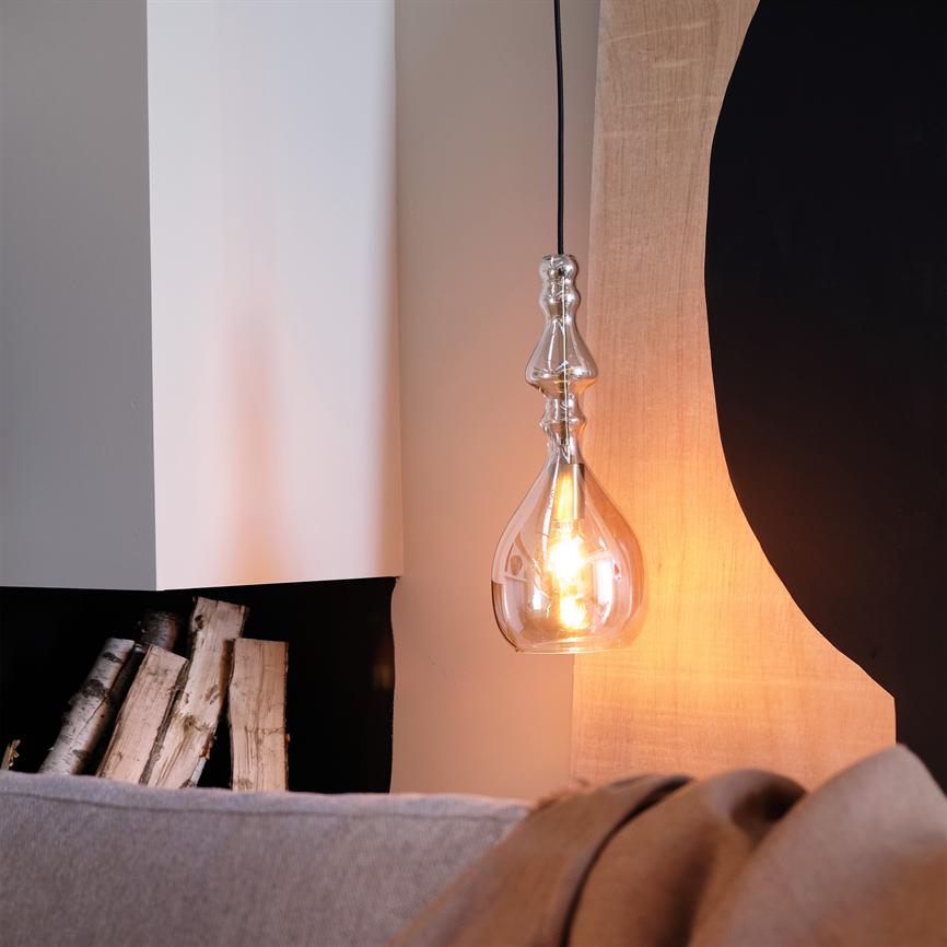 A decorative glass pendant light hangs and casts warm light; next to it are stacked birch logs in a niche, in front of a gray sofa and abstract wall decoration.