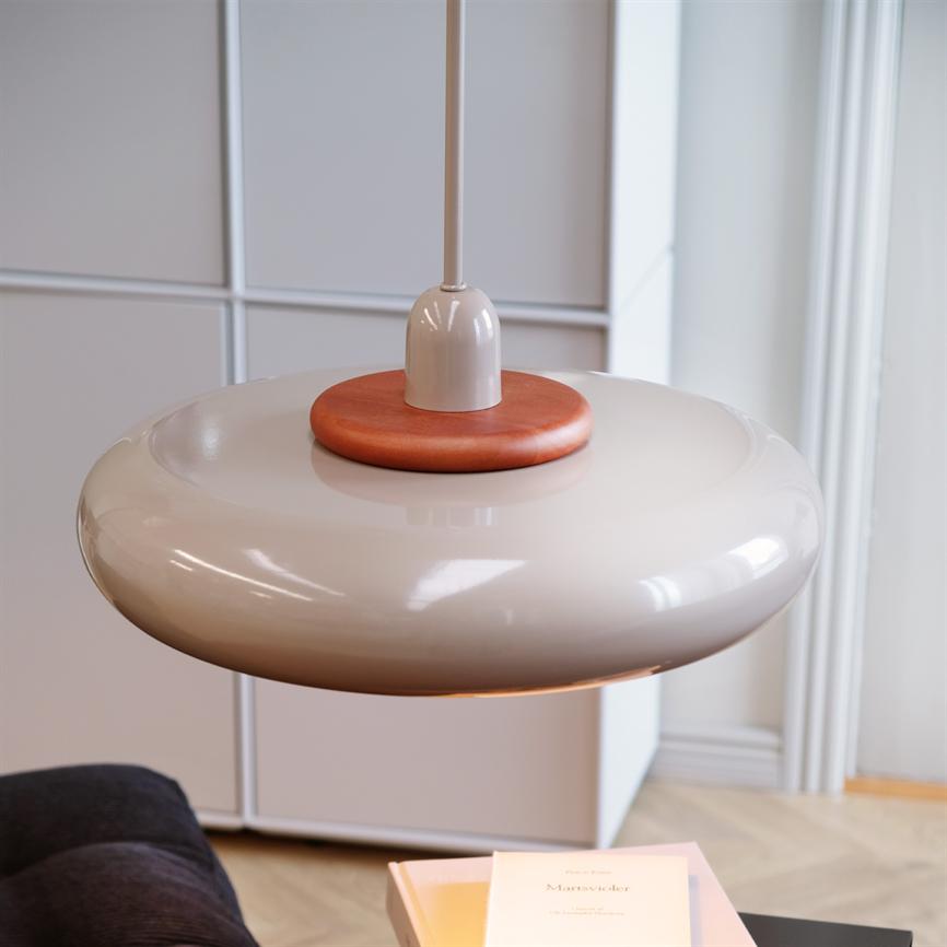 Pendant lamp hangs low over a table; glossy, round cream-coloured screen with wooden detail at the top, in a bright living room in front of cupboard and sofa. There is text on the book cover, but it is indistinct and illegible.