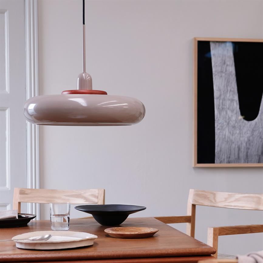 A round, beige pendant hangs over a wooden dining table; the table holds bowls, plates and a glass, surrounded by light wooden chairs and a framed abstract poster on the wall.