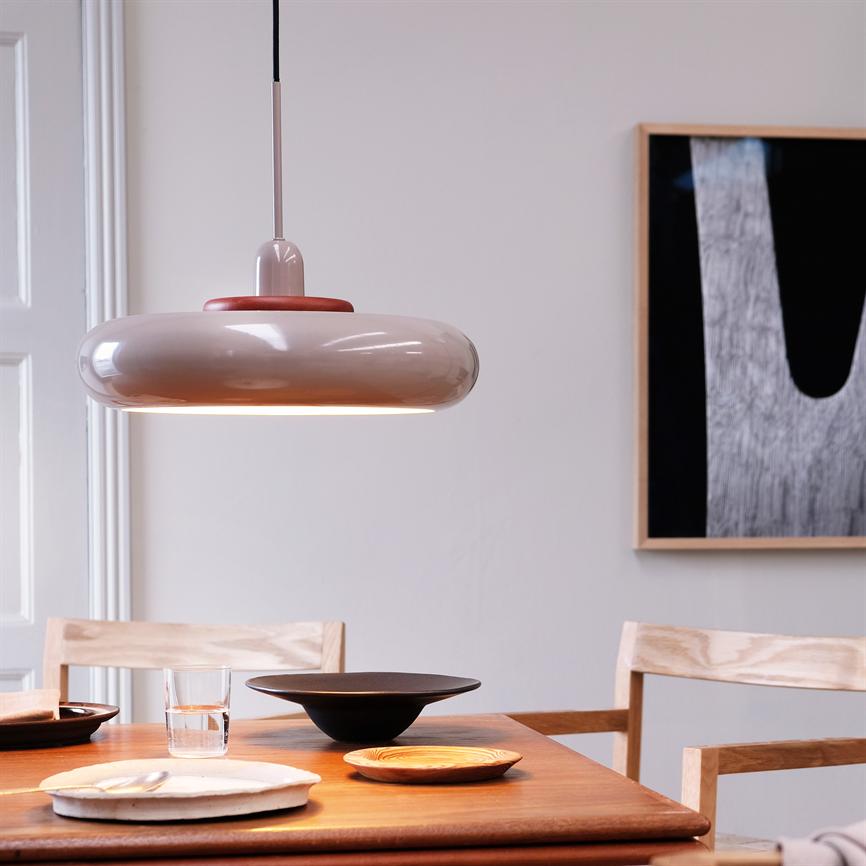 A round hanging lamp hangs above a wooden table and casts warm light on plates and a glass; surrounded by light wooden chairs and a simple wall decoration in a neutral space.
