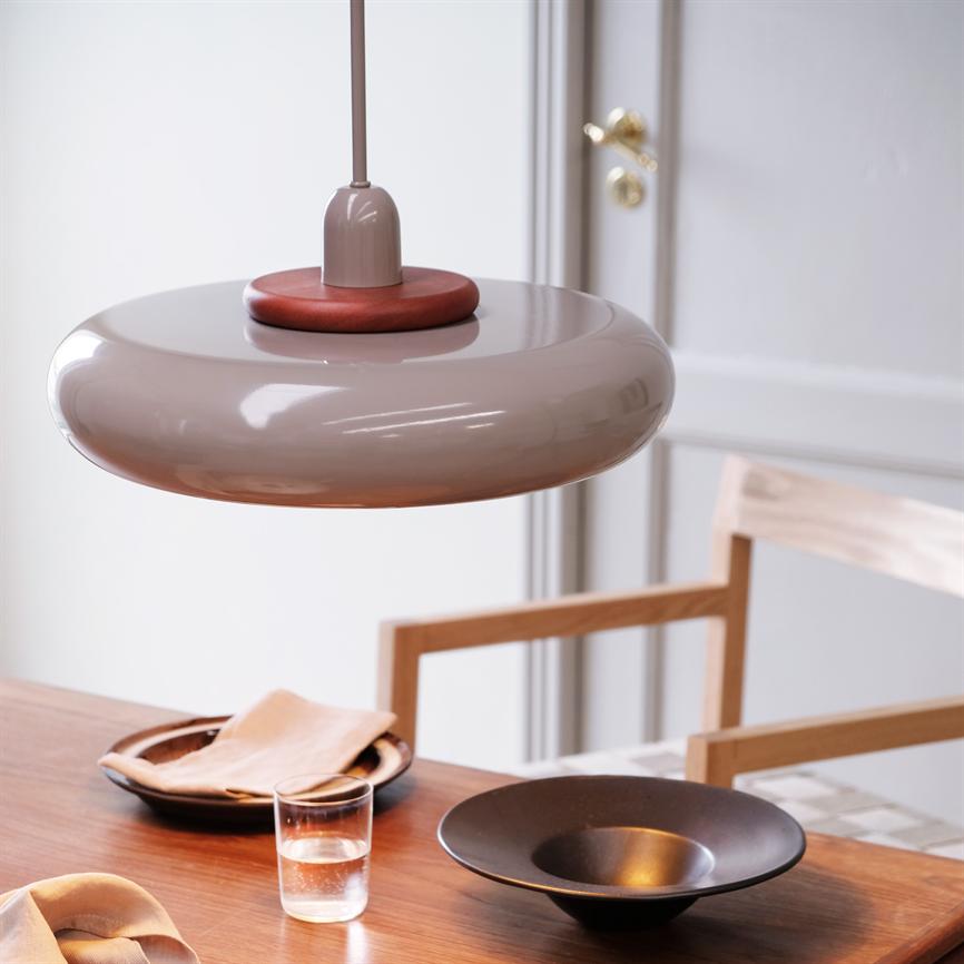 A round, beige pendant hangs over a wooden dining table; the table holds a black bowl, a plate with a napkin, a glass of water and a wooden chair in front of a white door.