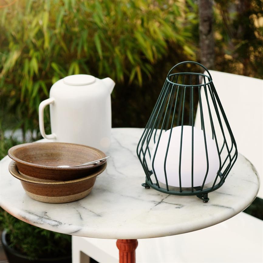 A green metal lantern with a white bulb sits on a round marble table, along with a white teapot and two brown bowls; outdoors, with green plants in the background.