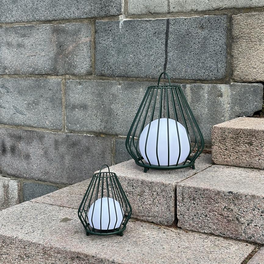 Two green-painted metal lanterns, one larger and one smaller, contain white globe lamps and stand on granite steps in front of a gray stone wall.