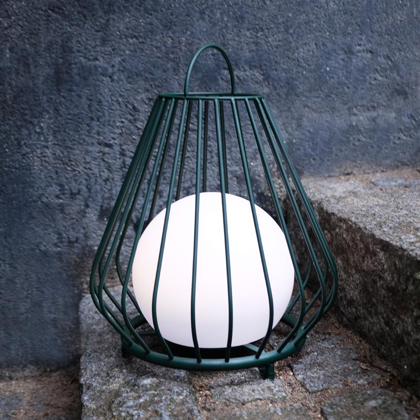 A spherical lamp glows inside a dark green metal cage, placed on rough granite steps outdoors in front of a gray, textured concrete wall.