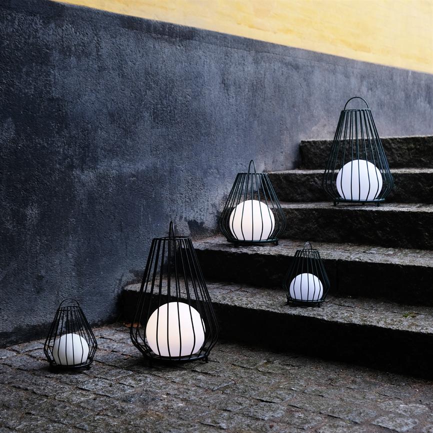 Black metal lanterns with white glowing balls are arranged and shine on an uneven granite staircase and cobblestones by a dark wall with a yellow stripe at the top.