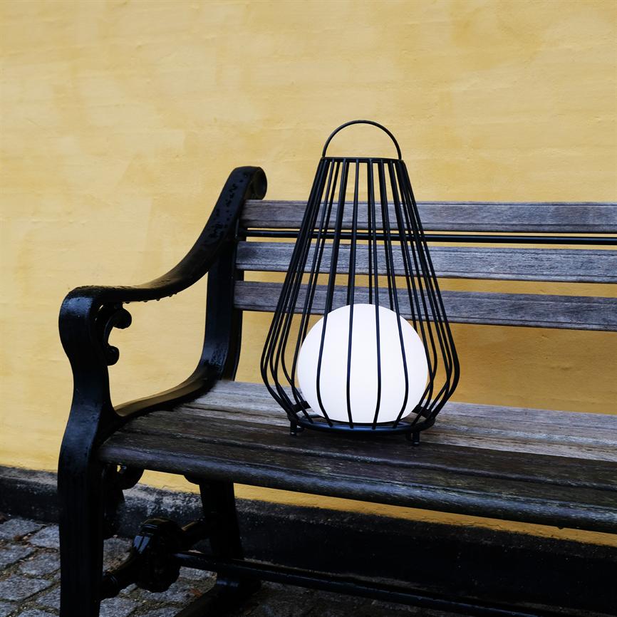 Black metal lamp with a white ball is placed on the seat of a worn wooden bench; the bench stands in front of a yellow wall above a cobbled pavement.