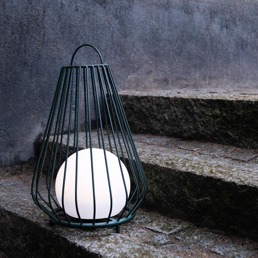A white spherical lamp in a dark green metal grid glows softly and stands on moss-covered, weathered stone steps in front of a gray, rough wall.