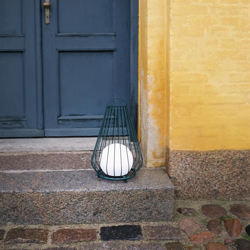 A green metal lantern with a white ball stands on a granite step in front of a blue door and a yellow plastered wall in a cobbled entryway.