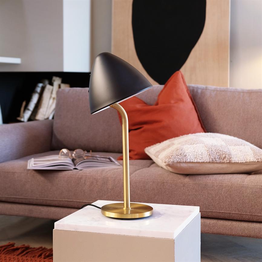 Table lamp with black shade and gold-coloured stand tilts the shade downwards and stands on a white marble block in front of a pink sofa with red cushions and an open book in the living room.