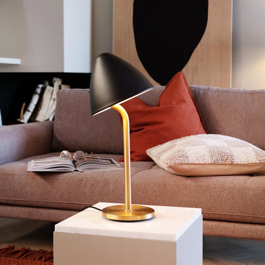 A table lamp with a black shade and golden base shines down on a white marble table; in front of a sofa with an open book, glasses and red and beige cushions in a cozy corner of the living room.