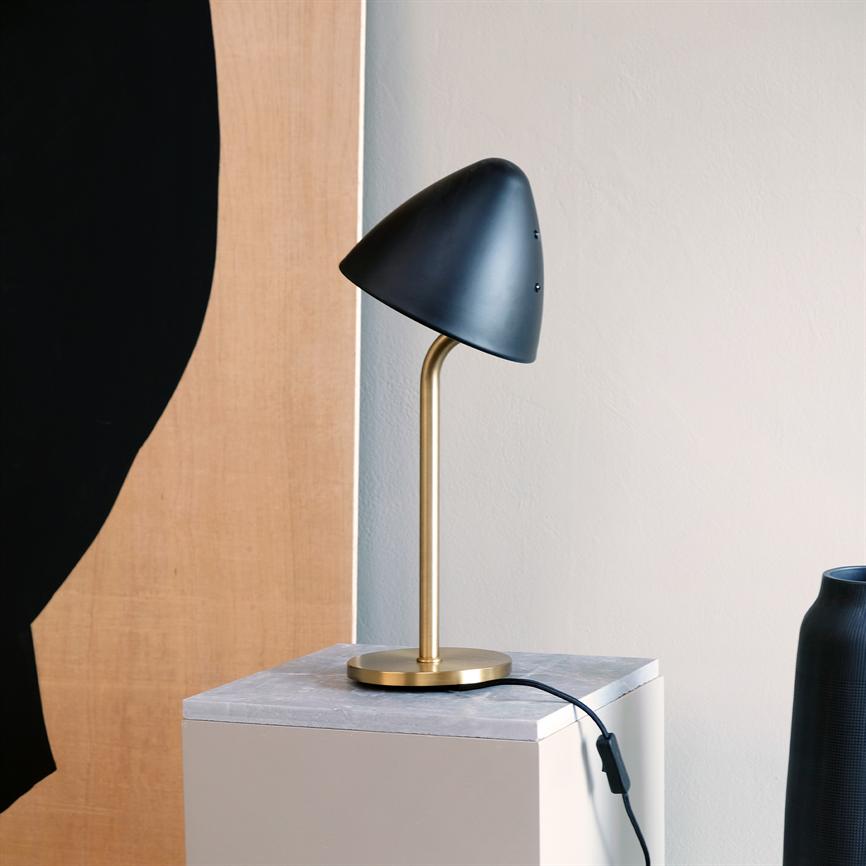 Table lamp stands on marble top on a box, black conical shade on curved brass rod, cord runs over edge; neutral wall, painting and dark vase in the background.