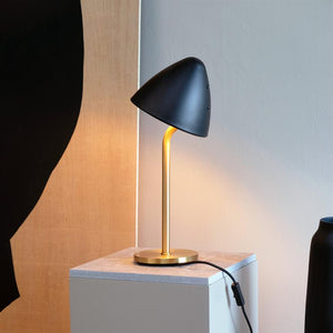 Black, mushroom-shaped table lamp emits warm light from a golden curved stand; it stands on a small square plinth in front of a light wall and wood paneling, with a black cord visible.