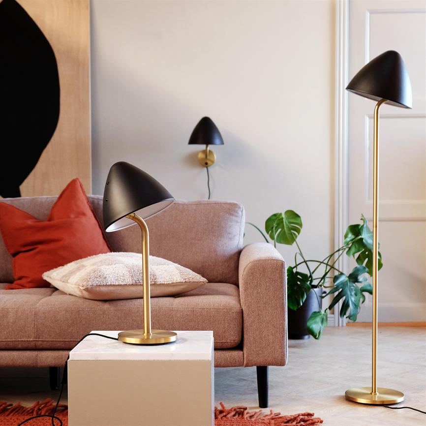 Black and brass table lamp shines down on a white side table; in front of a pink sofa with cushion cover and red pillow, next to a floor lamp and green houseplant.