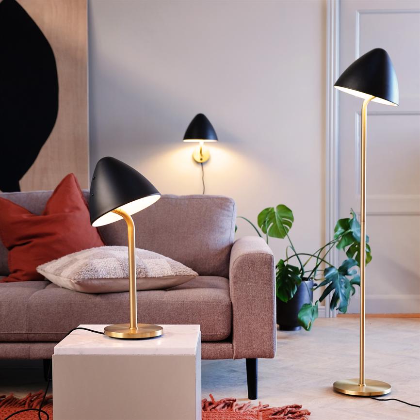 Three modern black and gold lamps cast soft, warm light over a light pink sofa in a cozy living room with cushions, a monstera plant and a small marble table.