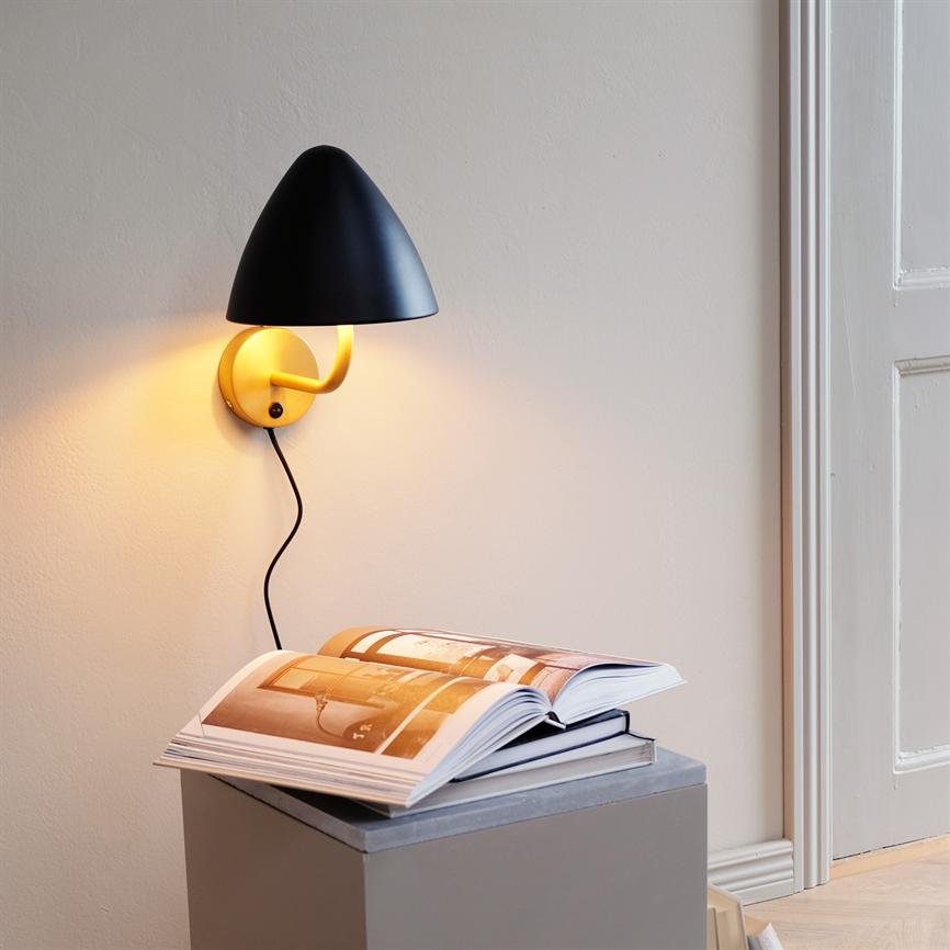 A wall lamp with a black shade and gold-coloured arm casts a warm light on an open picture book lying on a gray box by a white door in a calm, neutral room.