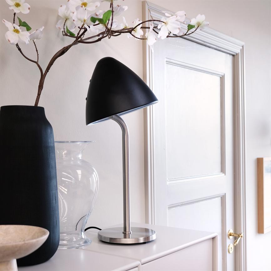 Table lamp points obliquely downwards, placed on a cabinet with a metal base and black shade, standing next to a black and a clear vase in front of a white door in a bright room.