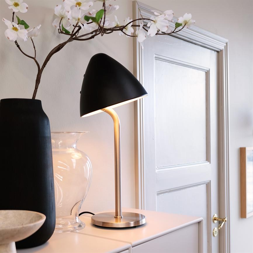 A black table lamp with a brass stem shines softly on a white cabinet, flanked by a tall black vase, a clear glass jar and a flowered branch in front of a white door.