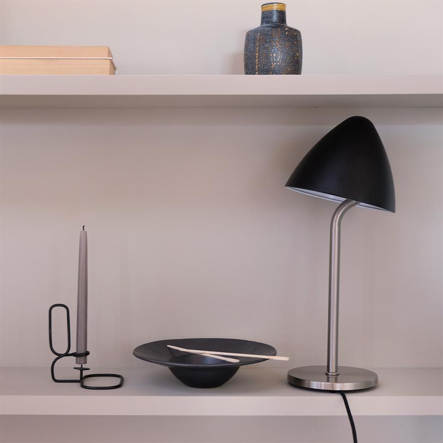 Black table lamp stands to the right on a light shelf next to a bowl of sticks and a candlestick; a vase and books are at the top against a neutral wall.