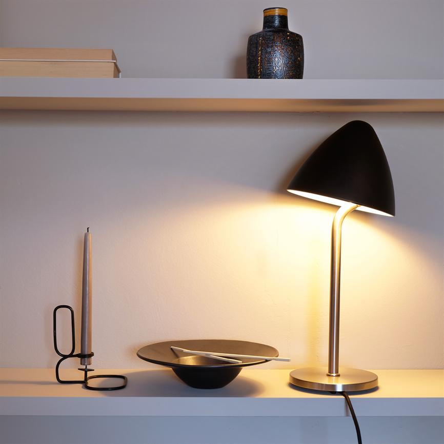 A black table lamp casts a warm light over a white shelf; next to it is a thin candlestick, a black bowl with cutlery, and a small decorative vase on the shelf above.
